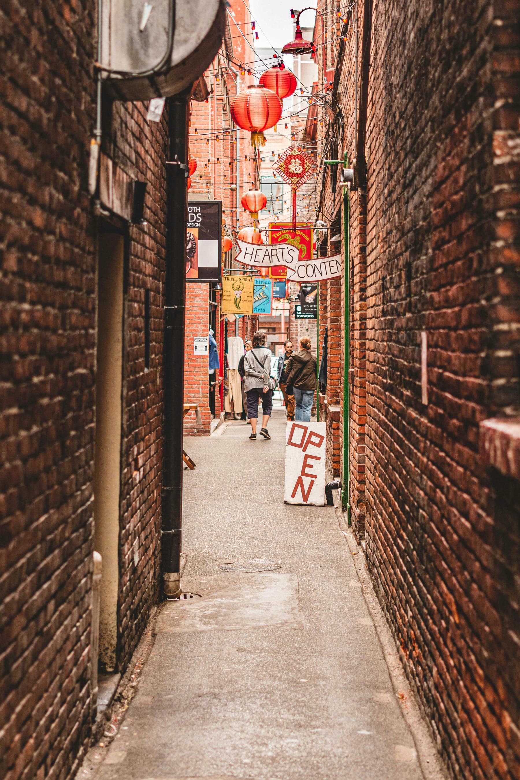 Narrow brick alley in Victoria, BC, strung with red lanterns and shop signs, with people strolling past an Open sign