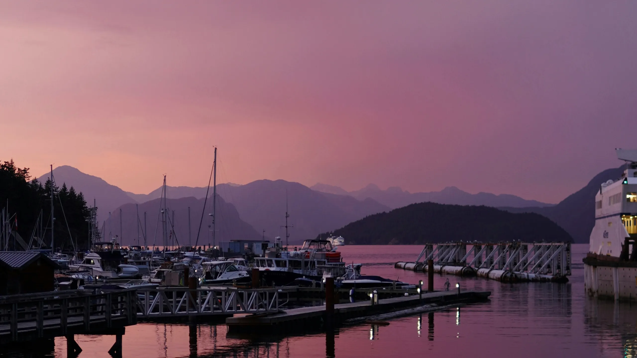 Marina at dusk under a pink sky, with boats at the docks and mountains across the water